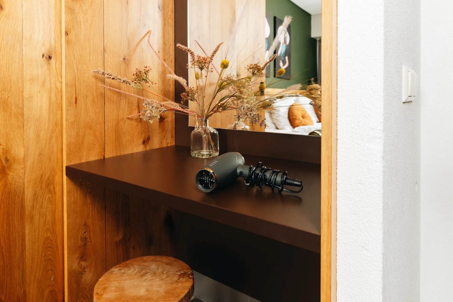 Stijlvolle kaptafel met een grote spiegel, een haardroger en een vaas met droogbloemen in een modern ingerichte hotelkamer.